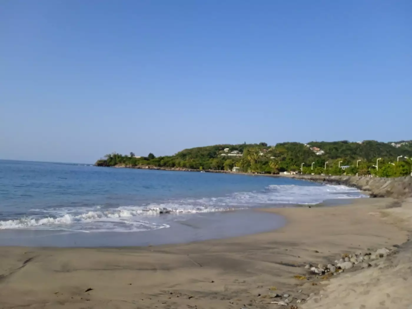 Sable gris et vaguelettes à l'anse Ferry, à Deshaies, avec la pointe Paul Thomas boisée en arrière-plan