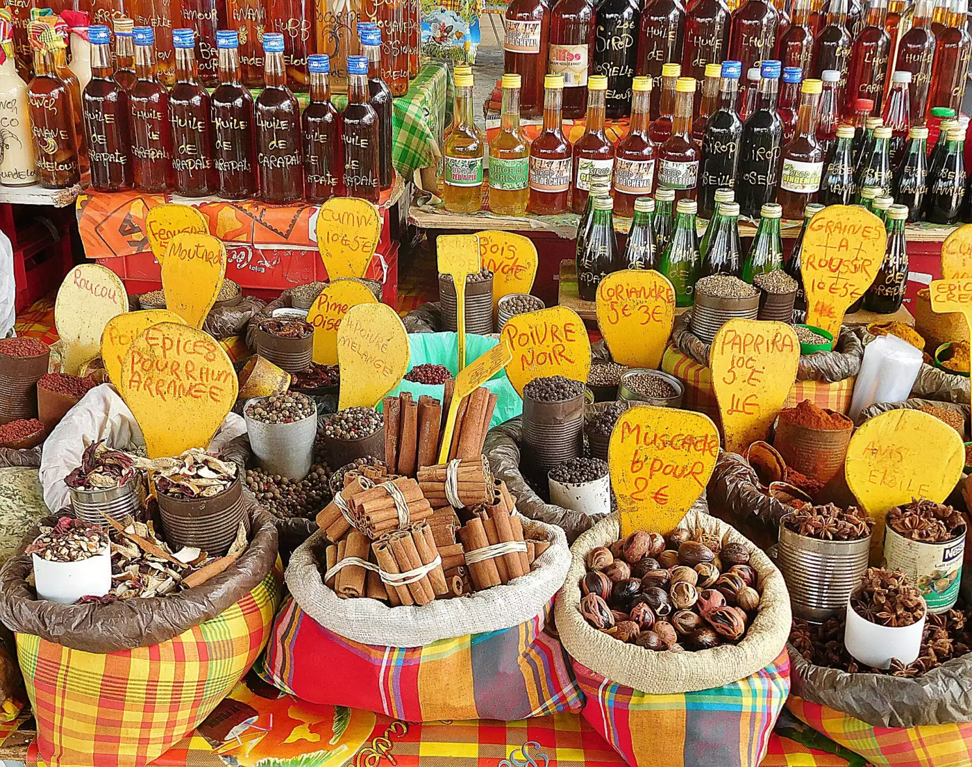 Étal du marché de la Darse, avec des sacs de cannelle, muscade, anis étoilé et paprika, des bouteilles de sirops et d'huile de carapate et des étiquettes de prix écrites à la main